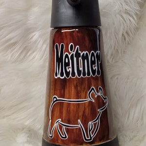 May include: A brown and black insulated water bottle with a wood grain design. The bottle has a black lid with a spout and a white pig silhouette on the side. The name "Meitner" is printed in white on the bottle.