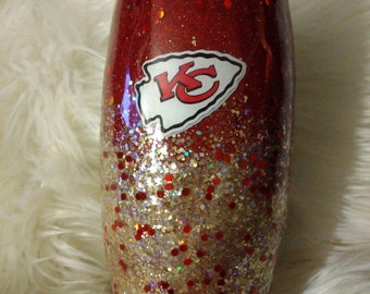 May include: A red and gold glitter tumbler with a white Kansas City Chiefs logo. The tumbler has a red glitter top and a gold glitter bottom.