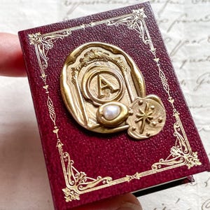 Puede incluir: Un pequeño libro de color burdeos con detalles dorados y un sello dorado con la letra "A" y una perla. El libro tiene un borde dorado decorativo y un pequeño dije en forma de estrella. El libro está en una mano.