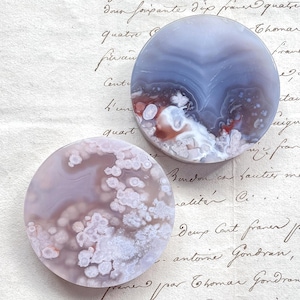 Puede incluir: Dos piedras de ágata redondas y pulidas con patrones y colores únicos. Una piedra presenta una mezcla de lavanda y blanco, la otra muestra tonos azules, blancos y rojos. Sobre un fondo de papel vintage.