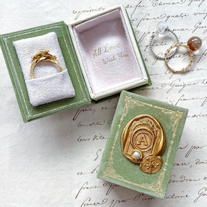 Op de afbeelding: Een gouden ring in een open groene fluwelen doos, naast een gesloten groene doos met een gouden zegel. De open doos heeft de tekst "All Love Wish You" binnenin. Twee andere ringen zijn zichtbaar.