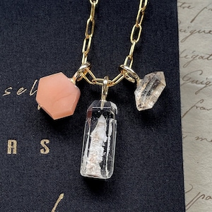 May include: A gold chain necklace with three crystal charms. The charms are a pink hexagonal crystal, a clear rectangular crystal, and a clear triangular crystal.
