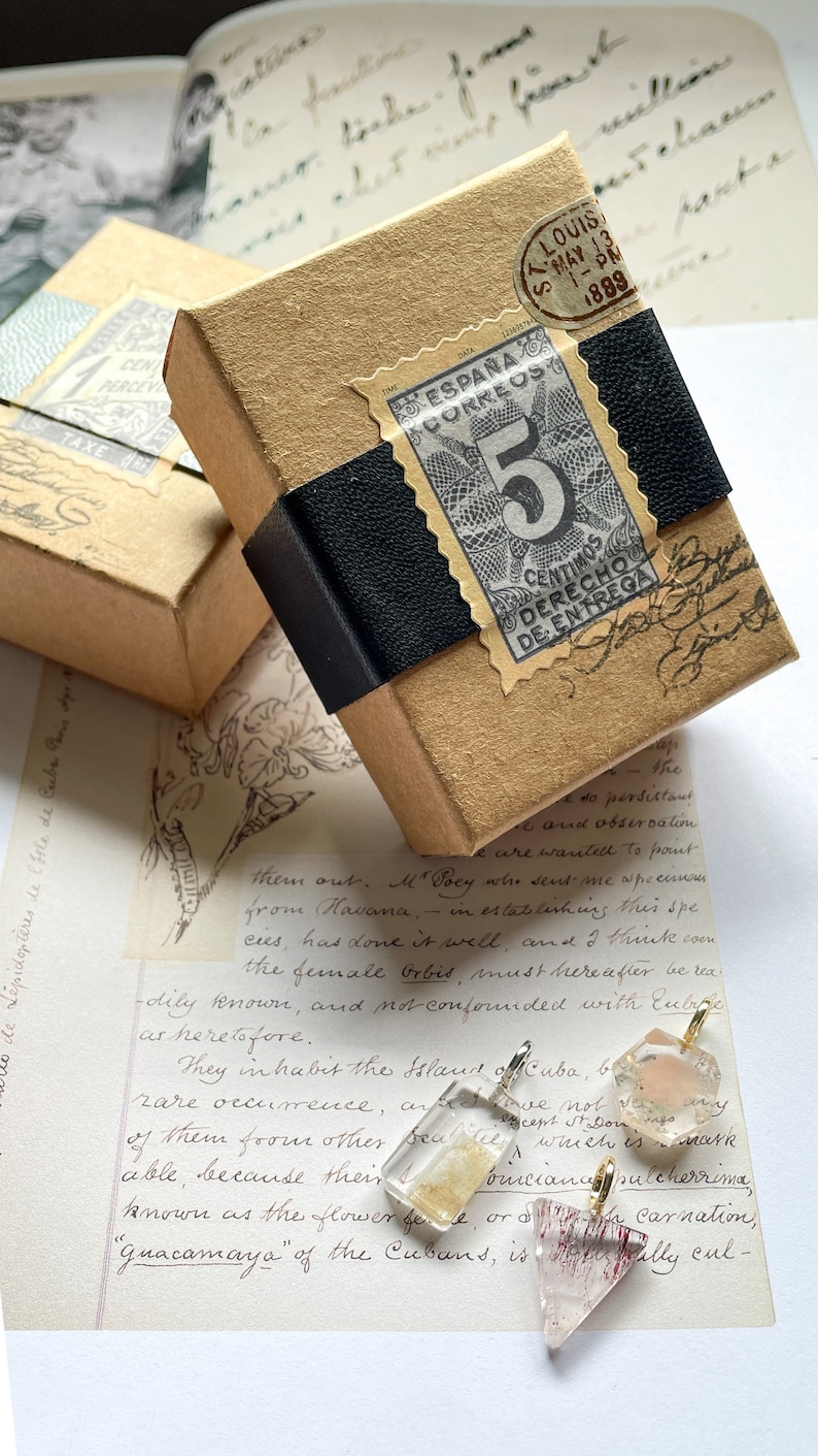 May include: Three clear glass pendants with gold accents and different colored crystals inside. The crystals are pink, gold, and red. The pendants are sitting on a piece of paper with handwritten text.