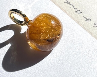 May include: A round, golden rutilated quartz pendant featuring visible needle-like inclusions within the translucent stone. The gemstone is securely set in a polished gold-tone metal bail, designed for attachment to a necklace chain or cord.