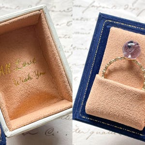May include: A gold ring with a purple gemstone is displayed in a blue velvet box with a peach-colored interior. The box is open, revealing the ring and the inscription "All Love Wish You" in gold lettering.