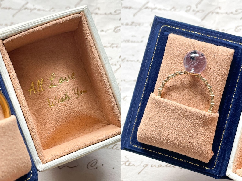 May include: A gold ring with a purple gemstone is displayed in a blue velvet box with a peach-colored interior. The box is open, revealing the ring and the inscription "All Love Wish You" in gold lettering.