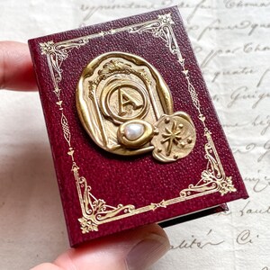 May include: A small, burgundy-colored book with gold filigree and a gold seal featuring the letter "A" and a pearl. A gold star-shaped emblem is also visible. The book is held in a person's hand.