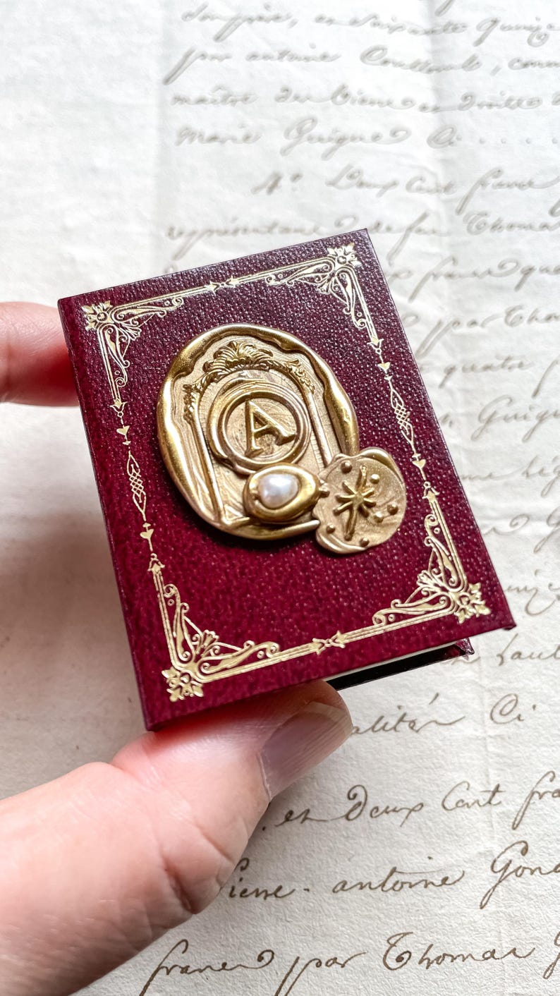 May include: A small, burgundy-colored book with gold filigree and a gold seal featuring the letter "A" and a pearl. A gold star-shaped emblem is also visible. The book is held in a person's hand.
