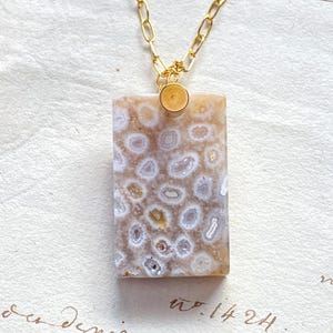 May include: A rectangular pendant necklace with a gold chain. The pendant is a light brown stone with white and tan circular patterns. The chain is a delicate gold link style. The background is a cream-colored paper with handwritten script.