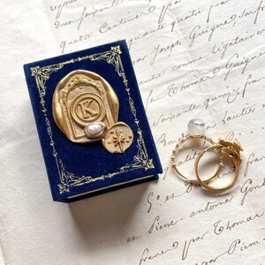 May include: A navy blue velvet jewelry box with gold embellishments and a gold seal featuring the letter "K" and a pearl. Two gold rings are next to the box, one with a floral design.
