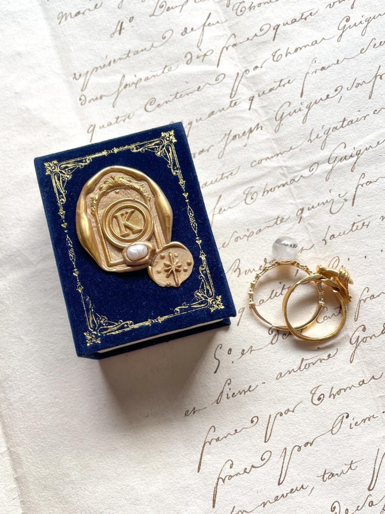 May include: A navy blue velvet jewelry box with gold embellishments and a gold seal featuring the letter "K" and a pearl. Two gold rings are next to the box, one with a floral design.