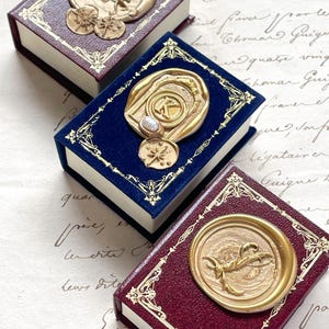 May include: Three miniature books with ornate gold-colored embellishments. The books have burgundy and navy blue covers with gold foil borders. Each book features a unique gold wax seal design, including celestial and initial motifs.