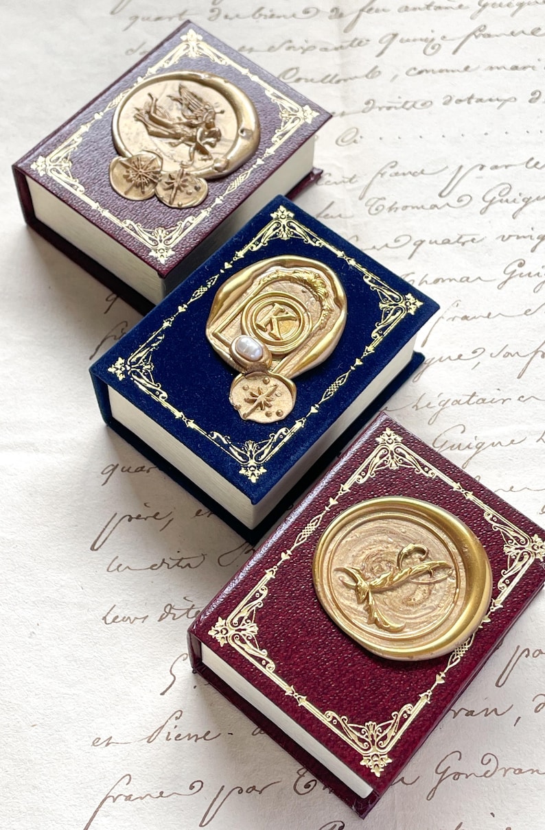 May include: Three miniature books with ornate gold-colored embellishments. The books have burgundy and navy blue covers with gold foil borders. Each book features a unique gold wax seal design, including celestial and initial motifs.