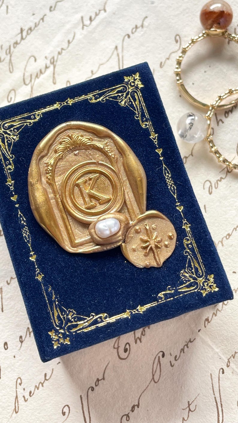 May include: A gold-colored wax seal with the letter "K" and a pearl accent, resting on a dark blue velvet box with gold trim. A smaller, starburst-shaped seal is next to it. Gold rings with gemstones are in the background.