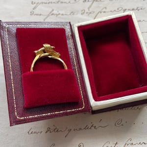 May include: A gold ring with an ornate design, displayed in a red velvet-lined jewelry box with a red and gold exterior. The ring is the focal point, showcasing its intricate details. The box is open, revealing the ring's presentation.