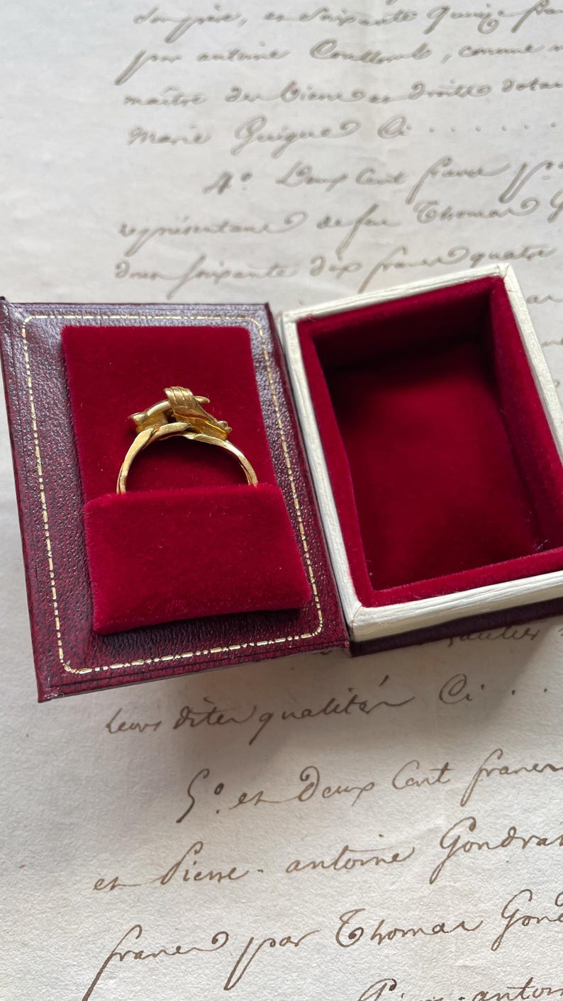 May include: A gold ring with an ornate design, displayed in a red velvet-lined jewelry box with a red and gold exterior. The ring is the focal point, showcasing its intricate details. The box is open, revealing the ring's presentation.