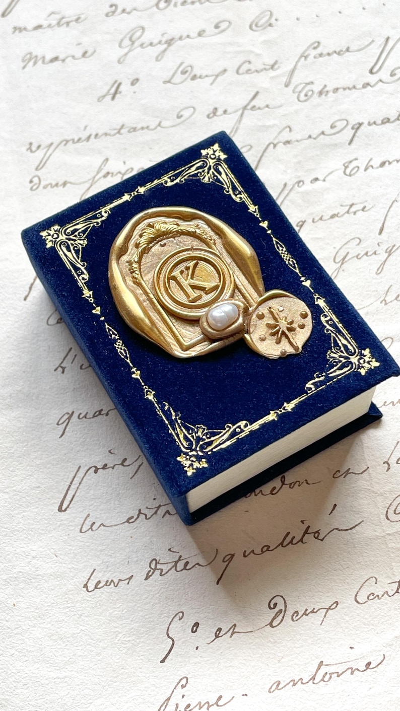 May include: A small, navy blue velvet book with gold filigree trim and a gold-colored seal. The seal features a letter "K" and a pearl. A compass-like design is also visible.