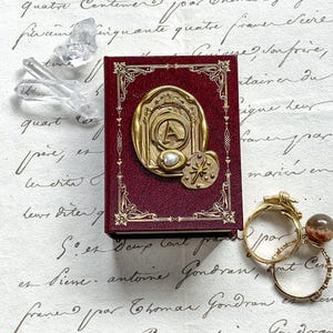 May include: A burgundy-colored miniature book with gold-colored embellishments and a large gold-colored emblem featuring the letter "A" and a pearl. The book is next to a few rings and crystals.