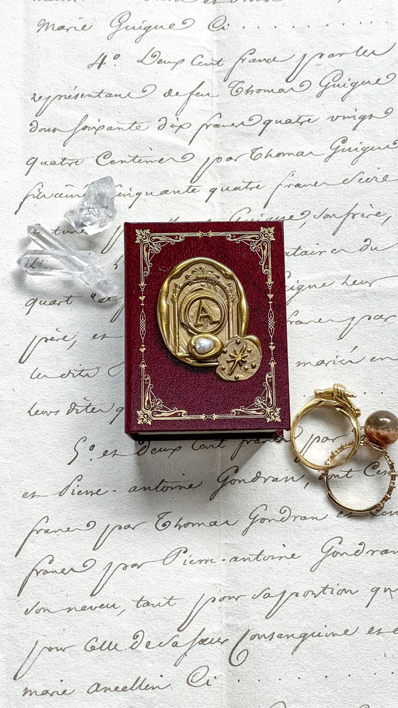 May include: A burgundy-colored miniature book with gold-colored embellishments and a large gold-colored emblem featuring the letter "A" and a pearl. The book is next to a few rings and crystals.