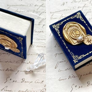 May include: A miniature blue velvet book with gold accents and a gold seal featuring the letter "K". The book has white pages and a decorative border. A small, decorative item is next to the book.