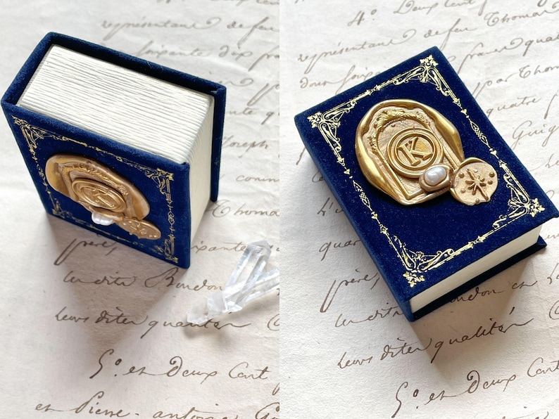 May include: A miniature blue velvet book with gold accents and a gold seal featuring the letter "K". The book has white pages and a decorative border. A small, decorative item is next to the book.