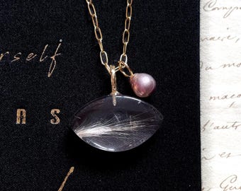 May include: A gold chain necklace featuring a marquise-shaped pendant containing fine white fibers encased in clear material, accompanied by a small, round, iridescent mauve pearl charm suspended from the same gold jump ring bail.