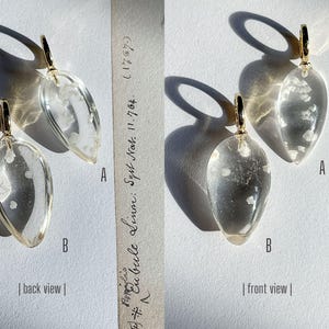 May include: A pair of clear glass earrings with a gold-tone metal hook. The earrings are shaped like teardrops and have a white flecked design. The earrings are shown from both the front and back views.