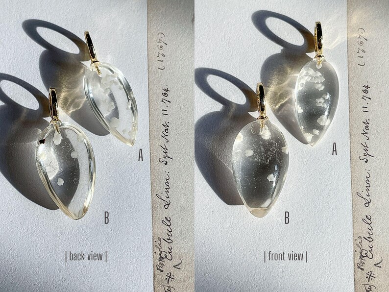 May include: A pair of clear glass earrings with a gold-tone metal hook. The earrings are shaped like teardrops and have a white flecked design. The earrings are shown from both the front and back views.