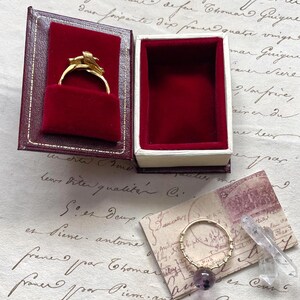 May include: A gold ring with a floral design inside a red velvet jewelry box. Beside it, a gold ring with a purple gemstone and a clear crystal. The background features handwritten script on aged paper.