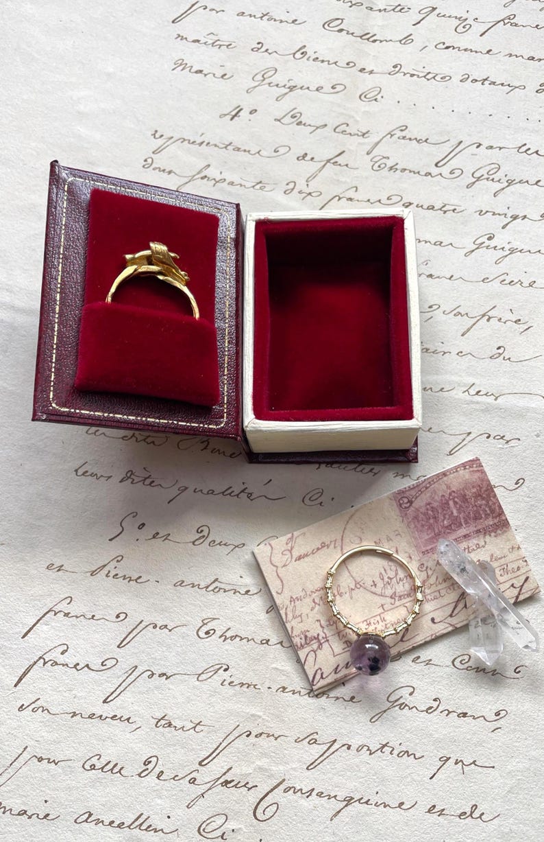 May include: A gold ring with a floral design inside a red velvet jewelry box. Beside it, a gold ring with a purple gemstone and a clear crystal. The background features handwritten script on aged paper.