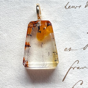 May include: A rectangular pendant with a gold bail. The pendant is clear with orange and black inclusions. The shape is wider at the top and tapers slightly towards the bottom. The background is a textured, off-white surface with handwritten script.