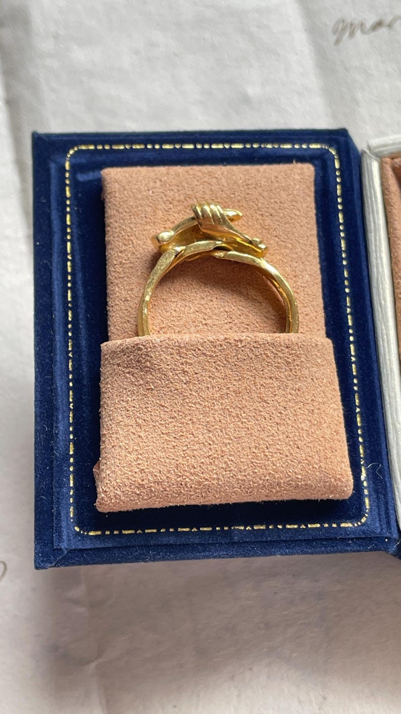May include: A gold ring with a unique design, presented in a blue velvet box with a tan interior. The ring features a detailed knot design, showcasing the craftsmanship and elegance of the jewelry.