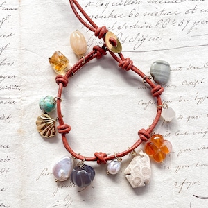 May include: A red leather bracelet adorned with diverse charms, such as gemstones, pearls, and a gold seashell. The bracelet is knotted and has a brass clasp. The background is vintage paper with handwritten script.