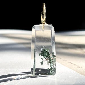 May include: A clear, rectangular pendant with a gold-coloured bail. The pendant contains green and white inclusions, creating a natural, organic appearance. The pendant is displayed on a white surface, with sunlight casting shadows.