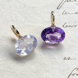 Puede incluir: Dos colgantes de piedras preciosas con aros dorados. Uno presenta una piedra azul claro de talla ovalada, y el otro una amatista morada oscura. Los colgantes se exhiben sobre una superficie texturizada blanquecina.