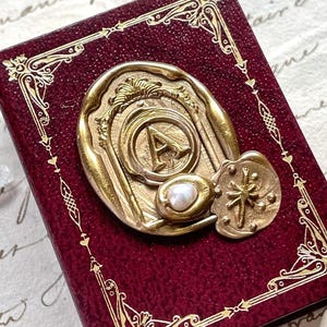 May include: A burgundy-colored, rectangular box with gold filigree edges and a gold seal. The seal features the letter "A" and a small pearl. The box has a vintage, antique aesthetic.