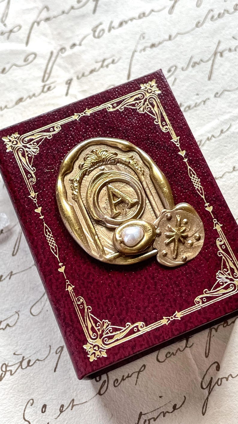 May include: A burgundy-colored, rectangular box with gold filigree edges and a gold seal. The seal features the letter "A" and a small pearl. The box has a vintage, antique aesthetic.
