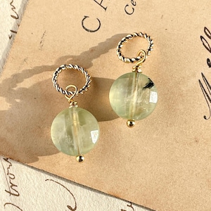Puede incluir: Un par de pendientes de piedra verde con aros de alambre dorado trenzado. Las piedras preciosas son redondas y tienen una apariencia translúcida.