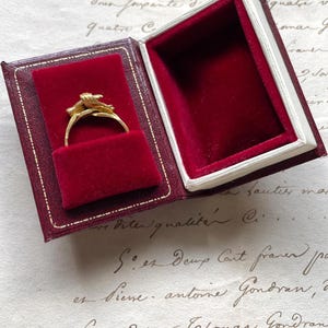 May include: A gold ring with a floral design is displayed in a burgundy velvet-lined box. The box is open, revealing the ring against the plush interior. The box has a white border and a burgundy exterior.