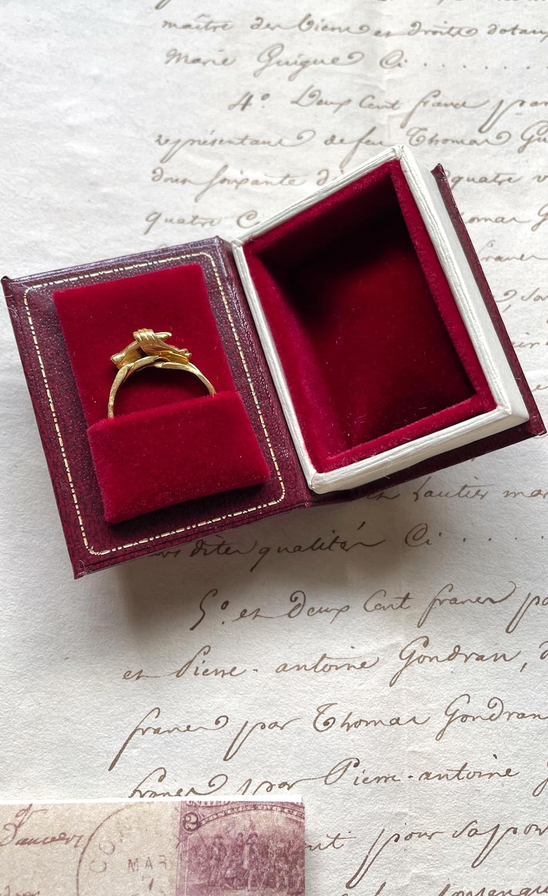 May include: A gold ring with a floral design is displayed in a burgundy velvet-lined box. The box is open, revealing the ring against the plush interior. The box has a white border and a burgundy exterior.