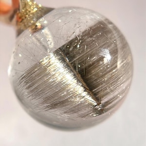 May include: A clear, spherical pendant with internal, silvery, needle-like inclusions. The pendant is attached to a gold-colored metal loop. The background is a soft, off-white color. The pendant is a unique piece of jewelry.