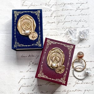May include: Two miniature books, one navy blue and one burgundy, with gold-colored embossed seals. The seals feature a letter, a pearl, and a compass rose design. The books are on a vintage paper background.