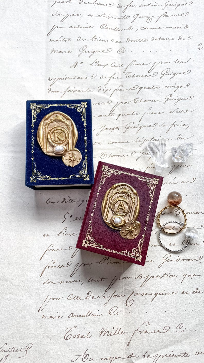 May include: Two miniature books, one navy blue and one burgundy, with gold-colored embossed seals. The seals feature a letter, a pearl, and a compass rose design. The books are on a vintage paper background.
