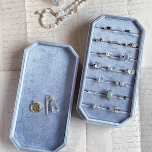 May include: Two light blue velvet jewelry trays, one containing a collection of various silver rings with gemstones and the other holding a gold coin pendant and two clear crystal charms, accompanied by a pearl necklace and small cosmetic containers.
