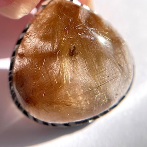 May include: A teardrop-shaped pendant with a silver bezel. The pendant features a translucent, golden-brown stone with delicate, hair-like inclusions. The stone is set against a bright white background, highlighting its unique texture and colour.