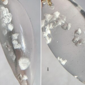 May include: Close-up of a clear, oval-shaped pendant with small, white, opaque flakes embedded inside. The pendant is hanging from a gold chain.