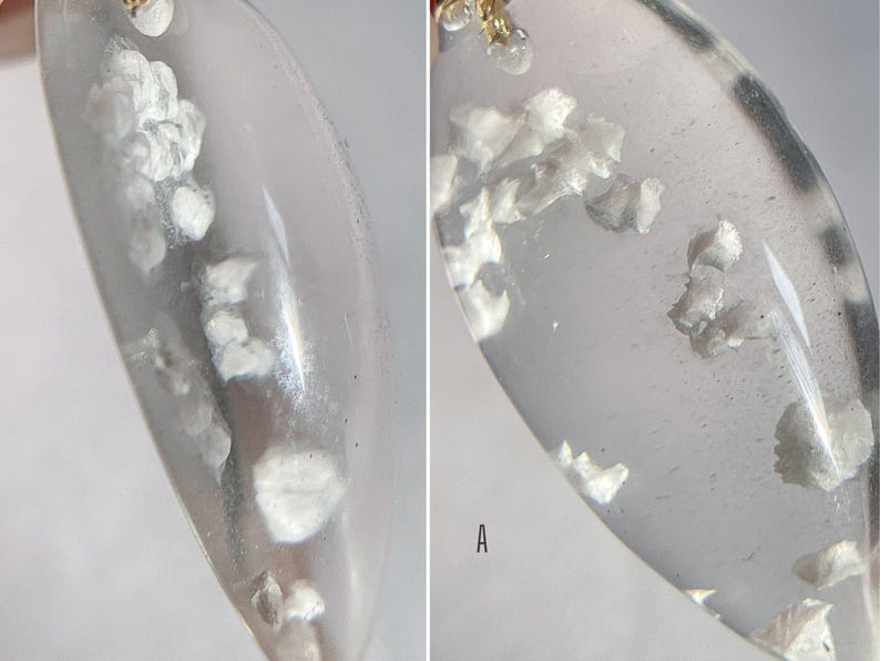 May include: Close-up of a clear, oval-shaped pendant with small, white, opaque flakes embedded inside. The pendant is hanging from a gold chain.
