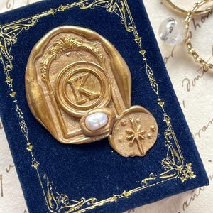 May include: A gold-colored wax seal with the letter "K" on a dark blue velvet box. The seal has a pearl and a starburst design. Gold rings with gemstones are in the background. The box has gold decorative trim.