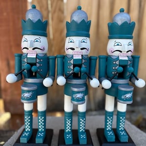 Puede incluir: Tres cascanueces de madera en color verde azulado y blanco, cada uno con una corona plateada y una cara pintada con bigote. Llevan uniformes y botas verde azulado con detalles blancos, sosteniendo baquetas y tambores con un logotipo de águila. Se apoyan en bases negras.