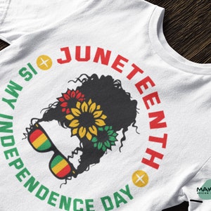 Puede incluir: Camiseta blanca con un gráfico circular verde, amarillo y rojo. El gráfico presenta una silueta negra de una mujer con el pelo rizado que lleva gafas de sol y una corona de flores. El texto "Juneteenth is my Independence Day" está escrito alrededor del círculo.